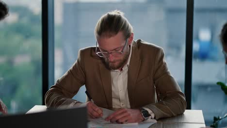 A-young-blond-man-with-glasses,-an-office-worker,-sits-at-a-table-in-a-light-suit-and-with-a-watch-on-his-hands-and-thinks-about-solving-issues.-Thinking-about-your-work-in-a-modern-office-with-a-panoramic-window