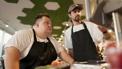 An-experienced-supermarket-worker,-a-man-with-a-beard-in-a-white-T-shirt-and-a-black-apron,-tells-an-overweight-male-trainee-how