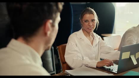 blonde-girl-with-eyeglasses-in-a-white-shirt-communicates-with-an-employee-in-a-white-shirt,-sitting-at-a-table-with-a-laptop-in-the-office