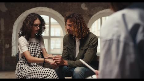 A-guy-with-curly-hair-in-a-green-jacket-and-a-brunette-girl-with-curly-hair-in-glasses-hold-hands-and-communicate-with-a-professional-psychologist,-a-girl-in-a-shirt,-in-a-brick-building