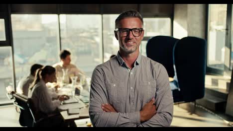 Successful-middle-aged-businessman-in-glasses-and-a-blue-shirt-folded-his-arms-on-his-chest,-smiling-and-looking-at-the-camera-in-an-office-with-large-windows