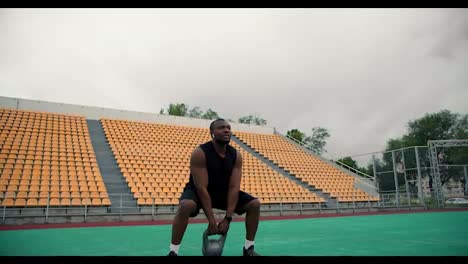 A-Black-man-in-black-sports-summer-clothes-is-training-using-a-kettlebell-in-a-stadium-with-yellow-chairs-in-the-stands
