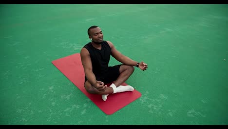 A-man-with-Black-skin-color-in-black-sports-summer-clothes-sits-on-a-red-mat-and-meditates-in-the-stadium