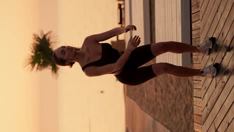 Vertical-video:-A-brunette-girl-with-tied-hair-in-a-black-sports-uniform-does-squats-on-a-sunny-beach-with-palm-trees,-a-beach-covered-with-boards