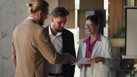 A-brunette-girl-in-round-glasses,-a-white-jacket-and-a-pink-shirt-communicates-with-her-colleagues-in-the-office,-a-blond-man-in-a-brown-jacket-and-a-guy-in-a-blue-jacket.-Discussing-important-ideas-at-work-in-a-fun-environment
