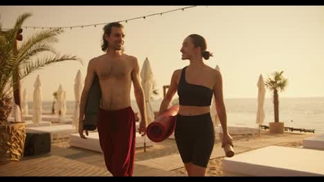 A-brunette-guy-in-red-pants-and-a-brunette-girl-in-a-black-sports-summer-uniform-are-walking-along-a-beach-covered-with-boards-and-talking-after-a-yoga-class