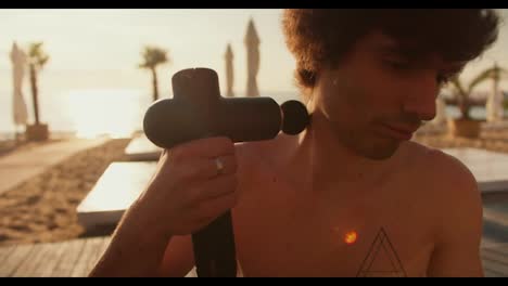 The-guy-massages-his-neck-with-a-special-vibrating-massager.-Relaxation-after-morning-workout-on-the-beach-at-sunrise