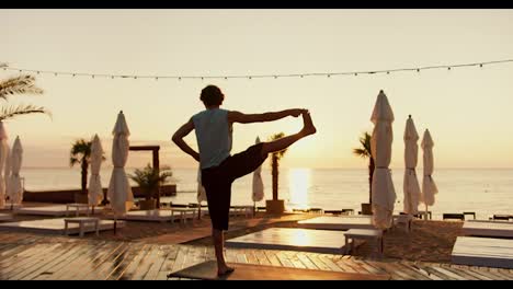 Rear-view-of-a-young-guy-who-raised-his-leg-up-and-stands-on-one-leg-on-a-sunny-beach-in-the-morning.-Yoga-and-stretching-classes-at-sunrise