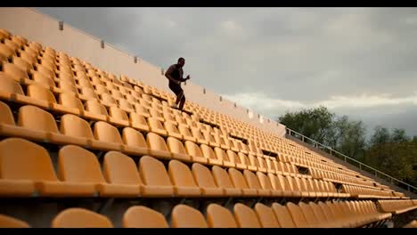 Athlete-with-Black-skin-color-in-a-black-sports-summer-uniform-and-white-bluetooth-headphones-walk-down-the-stadium-with-yellow-chairs-in-the-stands
