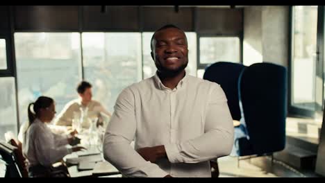 A-successful-businessman-with-Black-skin-in-a-white-shirt-folded-his-arms-on-his-chest,-smiles-and-looks-at-the-camera-against-the-backdrop-of-an-office-team-at-work