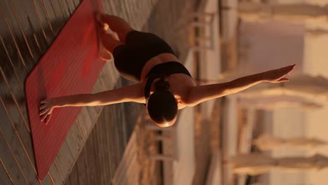 Vertical-video:-Rear-view-of-a-brunette-girl-in-a-black-sports-summer-uniform-doing-a-plank-on-one-arm-and-reaching-up-with-the-other.-Yoga-and-sports-class-on-a-sunny-beach-with-umbrellas-in-summer