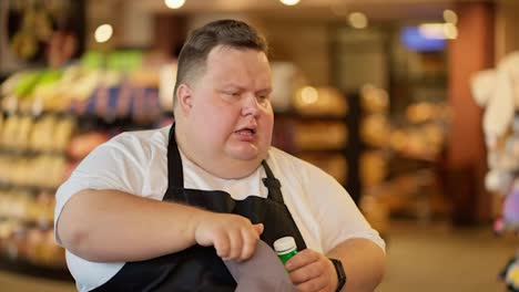 Tired-overweight-male-supermarket-worker-in-a-white-T-shirt-and-black-apron-drinks-water-from-a-green-bottle-and-wipes-sweat-from-his-forehead-during-a-break-during-a-hard-day-at-work