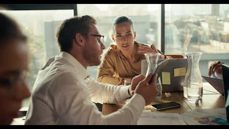An-office-worker-in-a-shirt-and-glasses-shows-his-project-on-a-tablet-to-a-blonde-woman,-sitting-at-a-table-in-an-office-with-panoramic-windows