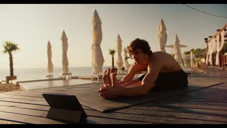 The-guy-does-the-stretch-following-the-video-on-the-tablet.-Online-stretching-and-yoga-classes.-Sunny-beach-in-the-morning