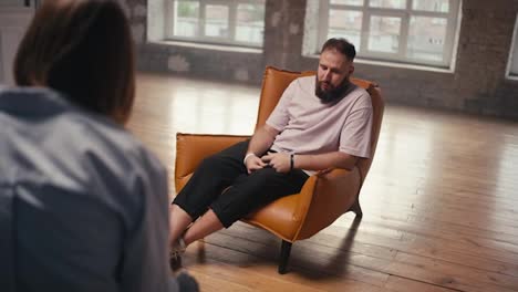 A-man-with-a-beard-in-a-light-pink-brown-chair-talks-about-his-problem-to-a-psychologist-at-group-therapy-in-a-brick-building