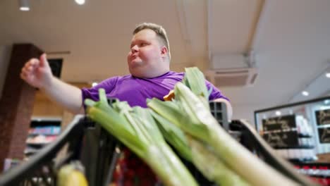 View-from-the-cart-side:-A-happy-overweight-man-wearing-a-purple-T-shirt-walks-through-the-supermarket,-looks-at-the-products.-Shallots-are-already-in-the-man's-cart