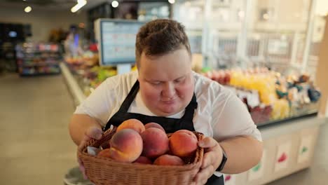Portrait:-An-overweight-man-at-a-supermarket-worker-in-a-white-T-shirt-and-a-black-apron-holds-a-basket-of-juicy-peaches-in-his-hands,-smells-them,-smiles-and-looks-at-the-camera