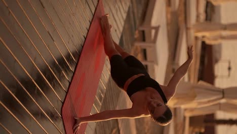 Vertical-video:-A-brunette-girl-in-a-black-sports-summer-uniform-does-a-plank-on-one-arm-and-stretches-up-with-the-other-on-a-sunny-beach-covered-with-boards.-Yoga-and-stretching-classes-during-golden-sunset-on-the-beach-in-summer