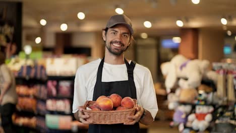 Portrait-of-a-happy-store-worker-in-a-white-T-shirt-and-a-black-apron,-a-man-holding-a-basket-of-peaches-in-his-hands-and-smiling