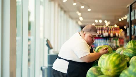 An-overweight-male-supermarket-worker,-a-man-in-a-white-T-shirt,-a-black-apron-and-a-gray-hat,-arranges-watermelons-and-checks-their-quality-on-the-supermarket-counter-while-eating-a-croissant
