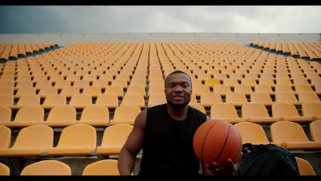 A-young-athlete-with-Black-skin-color-in-a-black-jersey-sits-on-the-yellow-stands-of-the-stadium-looks-at-the-camera-and-holds-a-basketball-in-his-hands