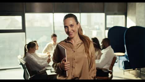 Happy-successful-businesswoman-in-business-brown-clothes-smiling-looking-at-the-camera-in-the-office.-Against-the-backdrop-of-office-workers