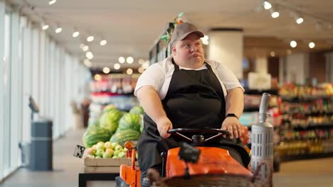 A-happy-overweight-supermarket-worker-in-a-white-T-shirt-and-a-black-apron-sits-on-a-special-cleaning-machine-in-a-cap-and-grimaces.-A-modern-cleaner-uses-a-machine-to-clean-the-floor-in-a-supermarket
