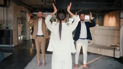 Group-yoga-class-in-a-modern-office.-A-girl-in-a-white-suit,-together-with-her-work-colleagues,-guys-in-brown-and-blue-jackets,-stand-in-a-special-pose-during-a-yoga-class-on-a-mat-in-a-modern-office