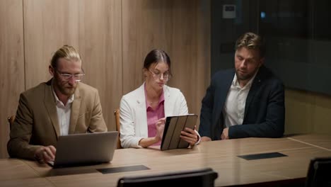 A-trio-of-office-workers-in-business-suits,-two-guys-and-a-girl,-discuss-solving-their-issues-using-a-laptop-and-an-electronic-tablet.-Business-meeting-of-office-workers-in-a-modern-office-with-wooden-flooring