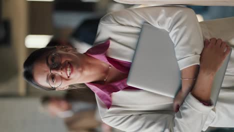 Vertical-video:-Portrait-of-a-brunette-girl-in-round-glasses,-white-jackets-and-a-pink-shirt-who-holds-a-laptop-in-her-hands-and-poses-against-the-background-of-a-modern-office-and-her-colleagues