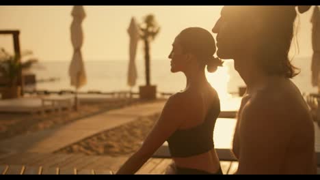 A-brunette-girl-in-a-black-sports-summer-uniform-is-meditating-with-a-guy-on-the-beach-by-the-sea-during-a-golden-sunrise