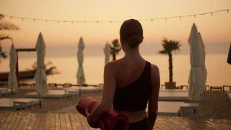 A-girl-in-a-black-summer-sports-uniform-with-a-mat-in-her-hands-goes-to-the-beach-to-do-morning-exercises-and-yoga.-Golden-colors-of-the-morning-beach-at-sunrise