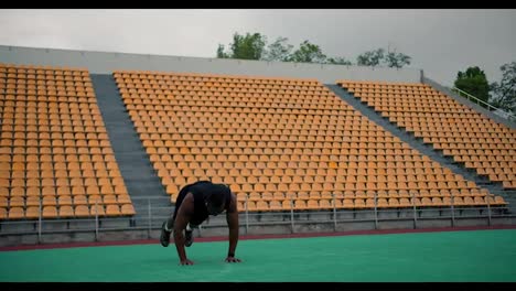Training-on-the-sports-field.-A-Black-man-in-black-sportswear-doing-a-burpee-exercise-in-a-yellow-stadium