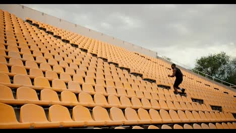 An-athlete-with-Black-skin-color-in-black-sports-summer-clothes-climbs-the-stairs-at-the-stadium-with-yellow-chairs-in-the-stands.-Sports-in-the-stadium