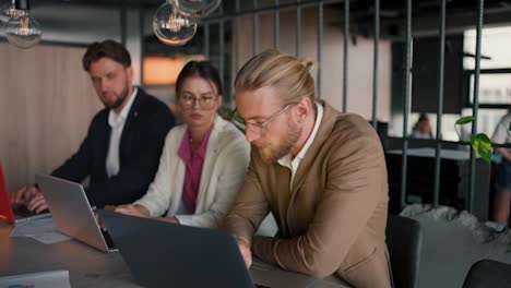 A-blond-man-in-glasses-with-a-beard-in-a-light-brown-suit-communicates-with-his-colleagues-at-the-table-while-caring-with-a-laptop-in-a-modern-office
