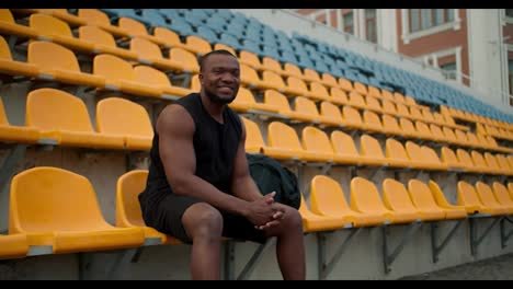 A-Black-skinned-athlete-in-a-black-sports-summer-uniform-sits-on-the-stands-of-the-stadium,-looks-at-the-camera-and-smiles