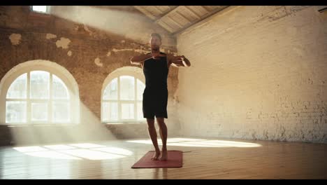 A-guy-in-black-clothes-does-exercises,-jumps-and-warms-up-on-a-special-rug-in-a-sunny-hall-with-white-brick-walls