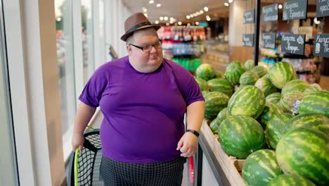 An-overweight-man-wearing-a-purple-T-shirt-and-a-brown-hat-walks-along-a-row-of-watermelons-in-a-large-supermarket-and-looks-at-them