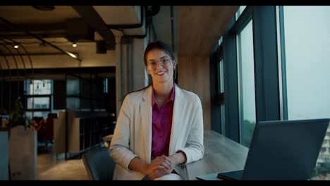 Portrait-of-a-brunette-girl-in-round-glasses,-a-white-jacket-and-a-pink-shirt-who-smiles-and-looks-at-the-camera-near-a-panoramic-window-in-a-modern-office