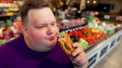 Close-up-shot-of-a-happy-overweight-man-with-short-hair-wearing-a-purple-T-shirt-eating-a-croissant-with-chocolate-filling-in-a-supermarket