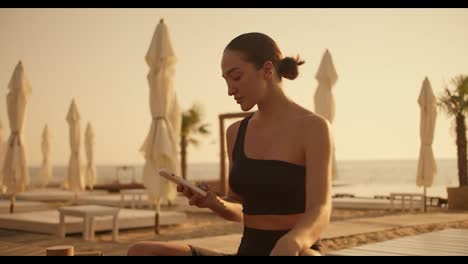 A-brunette-girl-in-a-black-top-works-on-a-smartphone-and-then-looks-away-on-a-sunny-beach-in-summer.-Golden-hour-on-the-beach