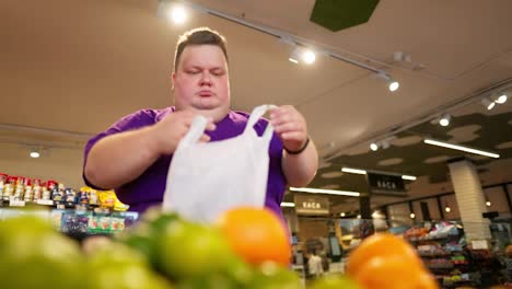 An-overweight-man-wearing-a-purple-T-shirt-chooses-oranges-from-the-counter-and-puts-them-in-a-bag-in-a-supermarket.-Daily-routine-of-an-overweight-man-in-a-supermarket.-Shopping-trip