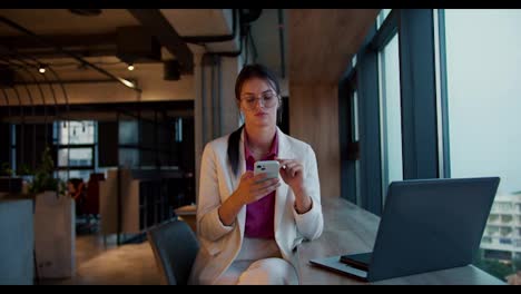 Portrait-of-a-brunette-girl-in-round-glasses-in-a-White-jacket-and-pink-shirt-who-works-in-a-White-telephone-near-a-panoramic-window-in-a-modern-office