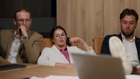 Tired-group-of-office-workers-leaning-on-the-back-of-their-chairs-and-sitting-at-a-table-in-a-modern-office.-A-brunette-girl-in-round-glasses-and-a-white-suit-sits-and-talks-with-two-guys-in-business-suits