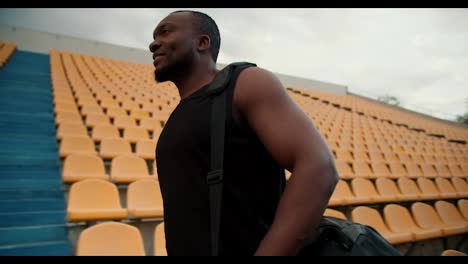 Overview-of-a-Black-skinned-athlete-in-a-black-T-shirt-with-a-bag-who-walks-through-the-stadium-with-yellow-chairs-in-the-stands