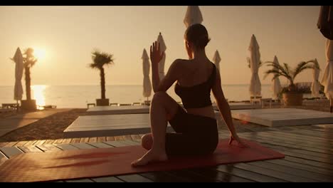 A-brunette-girl-in-a-black-sports-summer-uniform-does-yoga-on-a-red-mat-on-a-sunny-beach-in-the-morning.-Brunette-girl-doing-Spine-Twisting-Pose.-Golden-sunrise