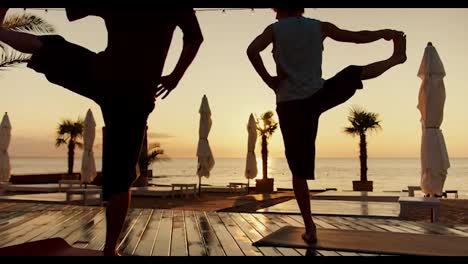 Two-guys-do-yoga-and-stand-on-one-leg-on-the-beach-in-the-morning.-Silhouettes-of-people-at-sunrise