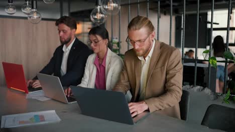 Portrait-of-a-young-blond-man-in-glasses-with-a-light-brown-beard-at-a-laptop-in-a-modern-office-with-his-colleagues