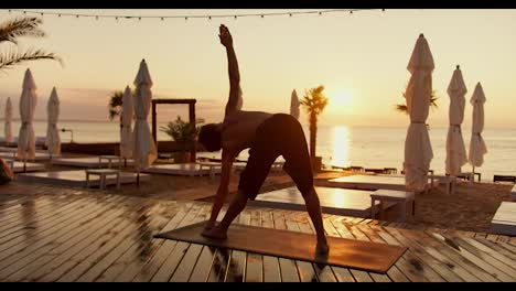 Ein-Junger-Mann-Macht-Yoga-Und-Nimmt-Morgens-Bei-Sonnenaufgang-An-Einem-Sonnigen-Strand-Verschiedene-Positionen-Seines-Körpers-Ein.-Wächst-An-Einem-Sonnigen,-Mit-Brettern-Bedeckten-Strand