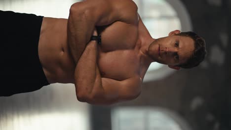 Vertical-video-portrait-of-a-muscular-male-athlete-with-a-naked-torso-who-poses-and-looks-at-the-camera-with-his-arms-folded-across-his-chest